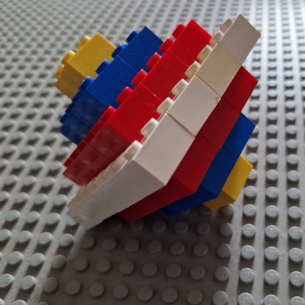 A shape made out of 2 by 2 lego bricks: the top half is a pyramid with a yellow, blue, red and white layer, and then the pyramid inverts back down to a point: red then blue then finally a single yellow square.